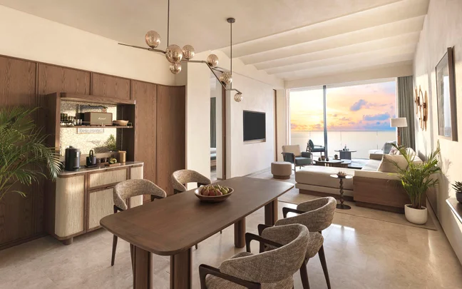 Dining and lounge space featuring natural wood furniture, indoor greenery, and expansive windows framing an ocean sunset. Fresh fruit bowl on the table enhances the setting.