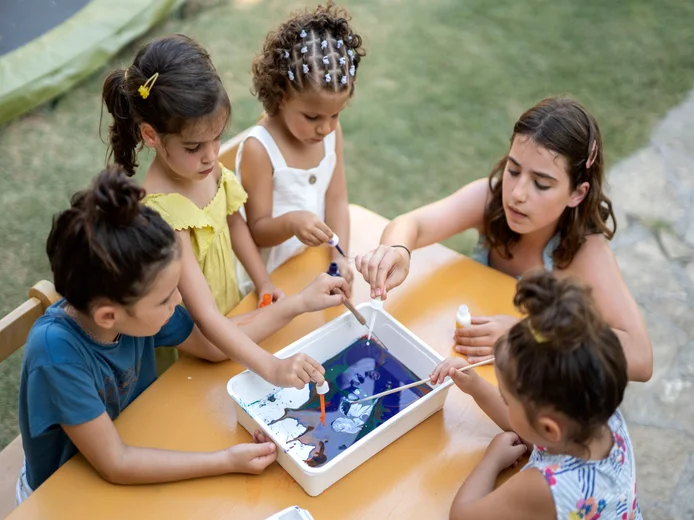 Children and a teen gather at an outdoor table, using droppers to mix colors in a water tray as part of an engaging art activity in natural sunlight.