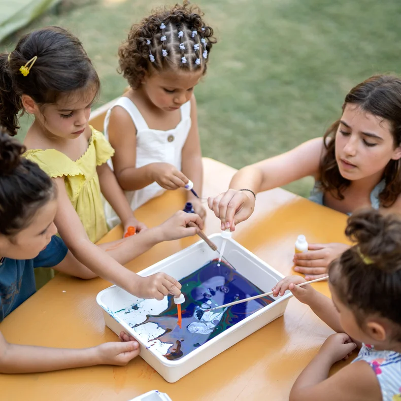 Children and a teen gather at an outdoor table, using droppers to mix colors in a water tray as part of an engaging art activity in natural sunlight.