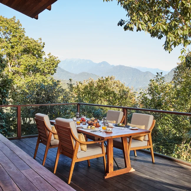 A patio features a dining table for four, framed by natural greenery and distant mountains beneath an open sky, offering a serene setting connected to nature.