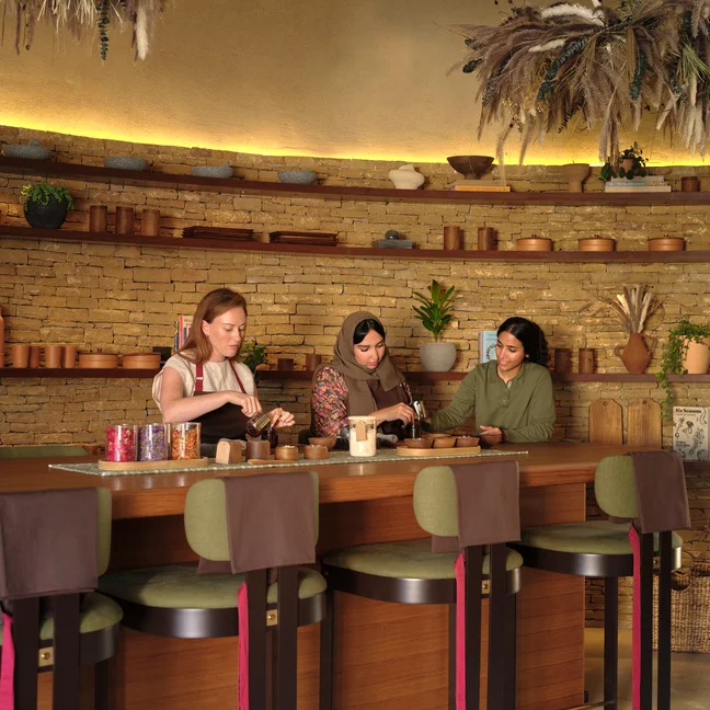 Guests craft together at a wooden bar in a warm space, featuring brick walls, greenery on shelves, and thoughtfully arranged dried flowers overhead.