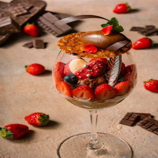 Glass with layered strawberries, blueberries, chocolate and wafers, set on a light surface with fresh strawberries and chocolate pieces arranged nearby.