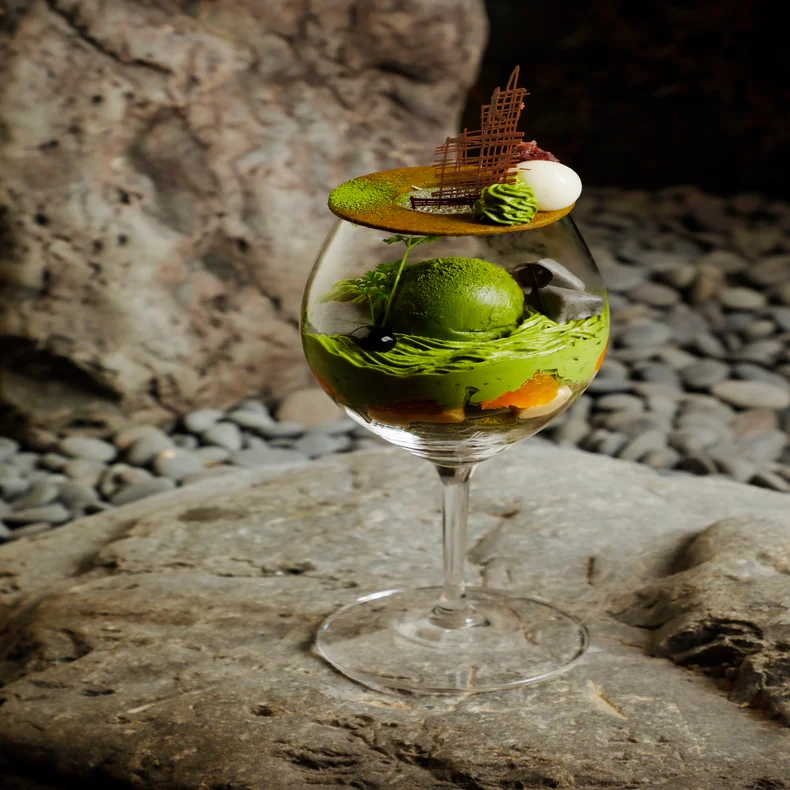 A glass with matcha dessert—layers of green ice cream, whipped cream, fruit, and chocolate—rests on a gray stone surface with pebbles in the background.