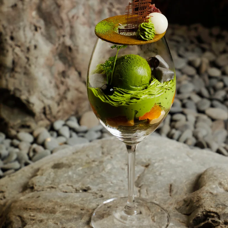 緑色のアイスクリーム、ホイップクリーム、フルーツ、チョコレートを重ねた抹茶のデザートを入れたグラスが、小石を背景にした灰色の石の上に置かれている。.