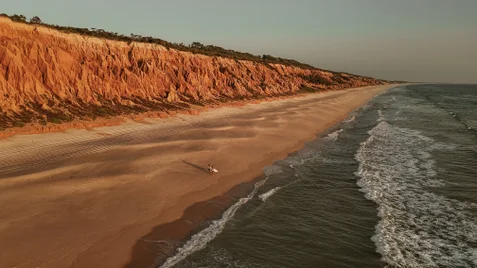 A solitary figure moves along a wide sandy beach beside gentle ocean waves, with tall orange cliffs bordering the shoreline and a clear sky overhead.