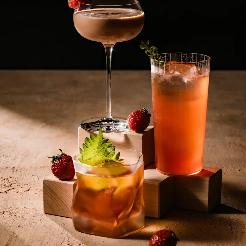 新鮮なフルーツを添えた3種類の飲み物が木製のブロックの上に置かれている。イチゴはその近くに置かれ、温かみのある茶色を背景に落ち着いた演出になっている。.