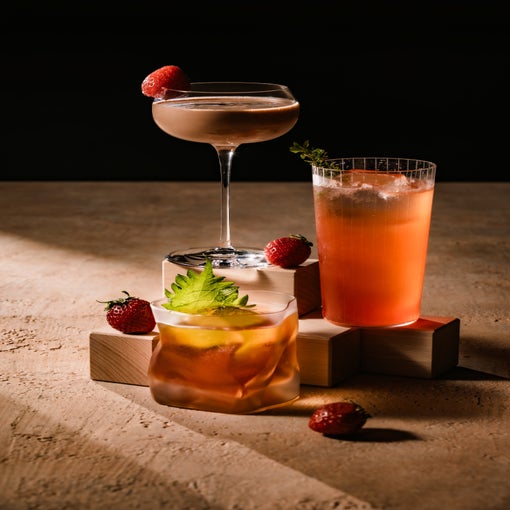 新鮮なフルーツを添えた3種類の飲み物が木製のブロックの上に置かれている。イチゴはその近くに置かれ、温かみのある茶色を背景に落ち着いた演出になっている。.