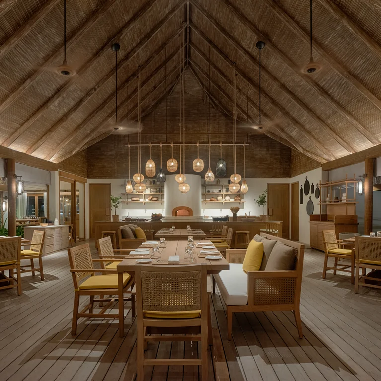 مطعم في الهواء الطلق مع سقف مرتفع من القش وطاولات وكراسي خشبية وإضاءة معلّقة وأدوات مائدة مختارة بعناية لتناول وجبة مريحة.