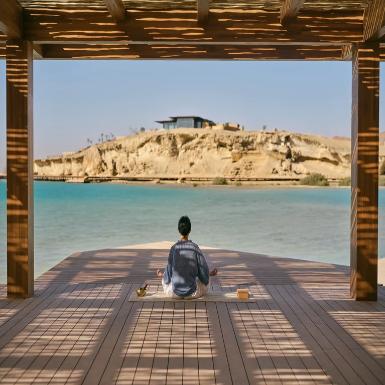 Individual practicing meditation on a wooden deck with views of serene blue water, rocky shoreline, and filtered sunlight beneath a shaded pergola.