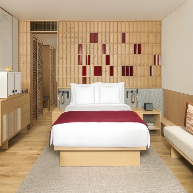 Spacious guest room with a large bed, crisp white linens accented in red, natural wood furnishings, built-in closet, and decorative wooden wall featuring subtle red details.