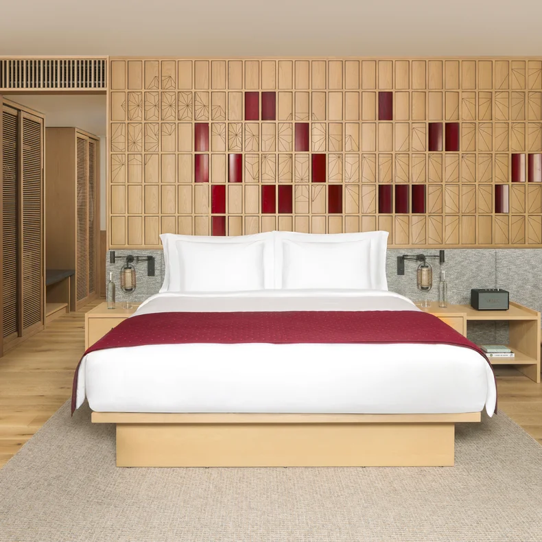 Spacious guest room with a large bed, crisp white linens accented in red, natural wood furnishings, built-in closet, and decorative wooden wall featuring subtle red details.