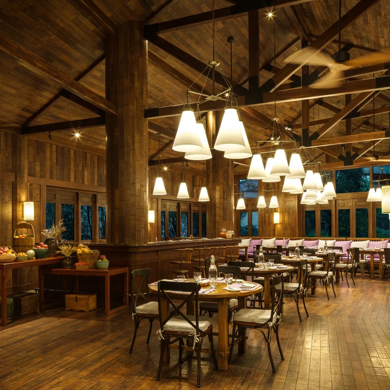 Restaurante con suelos, paredes y techo de madera natural. Mesas y sillas dispuestas cómodamente bajo modernas lámparas colgantes. El espacio está iluminado uniformemente.