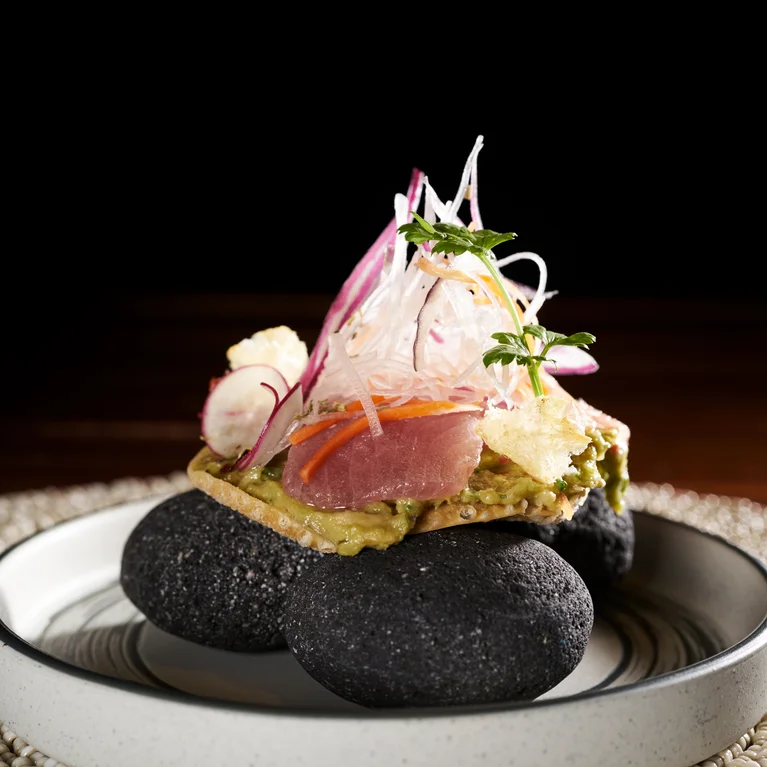 ترتكز التونة المشوية على قاعدة مقرمشة مع الأفوكادو وشرائح الفجل والخضراوات الدقيقة، مقدمة على حجارة سوداء ناعمة في وعاء أبيض.