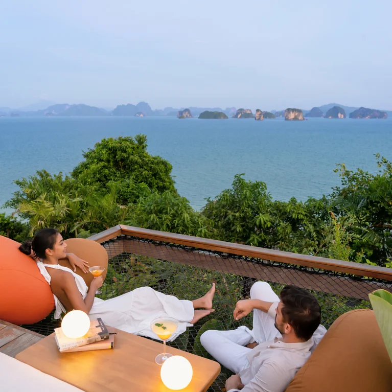 客人们坐在户外躺椅上,在郁郁葱葱的绿树中欣赏海景和岛屿风光,每人手持一杯饮料,尽情放松。.