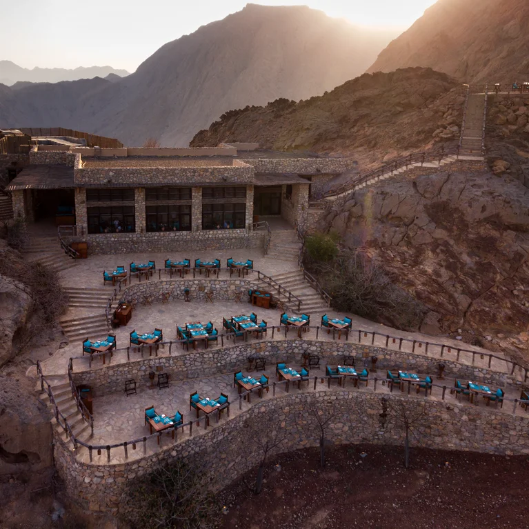 Comedor al aire libre con manteles azules, elevado en la ladera de una montaña al atardecer, con vistas al terreno natural y a los picos escalonados en la distancia.