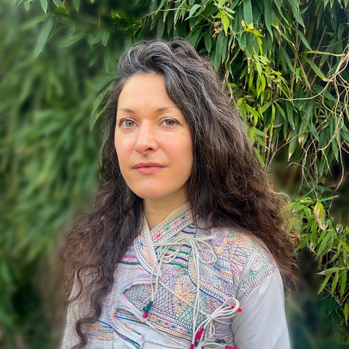 Individual with long wavy brown hair stands outside near lush greenery, dressed in a light embroidered top, reflecting natural surroundings and mindful design.
