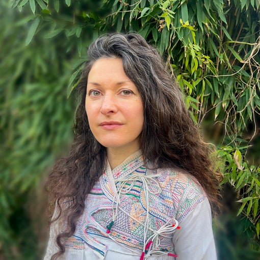 Individual with long wavy brown hair stands outside near lush greenery, dressed in a light embroidered top, reflecting natural surroundings and mindful design.