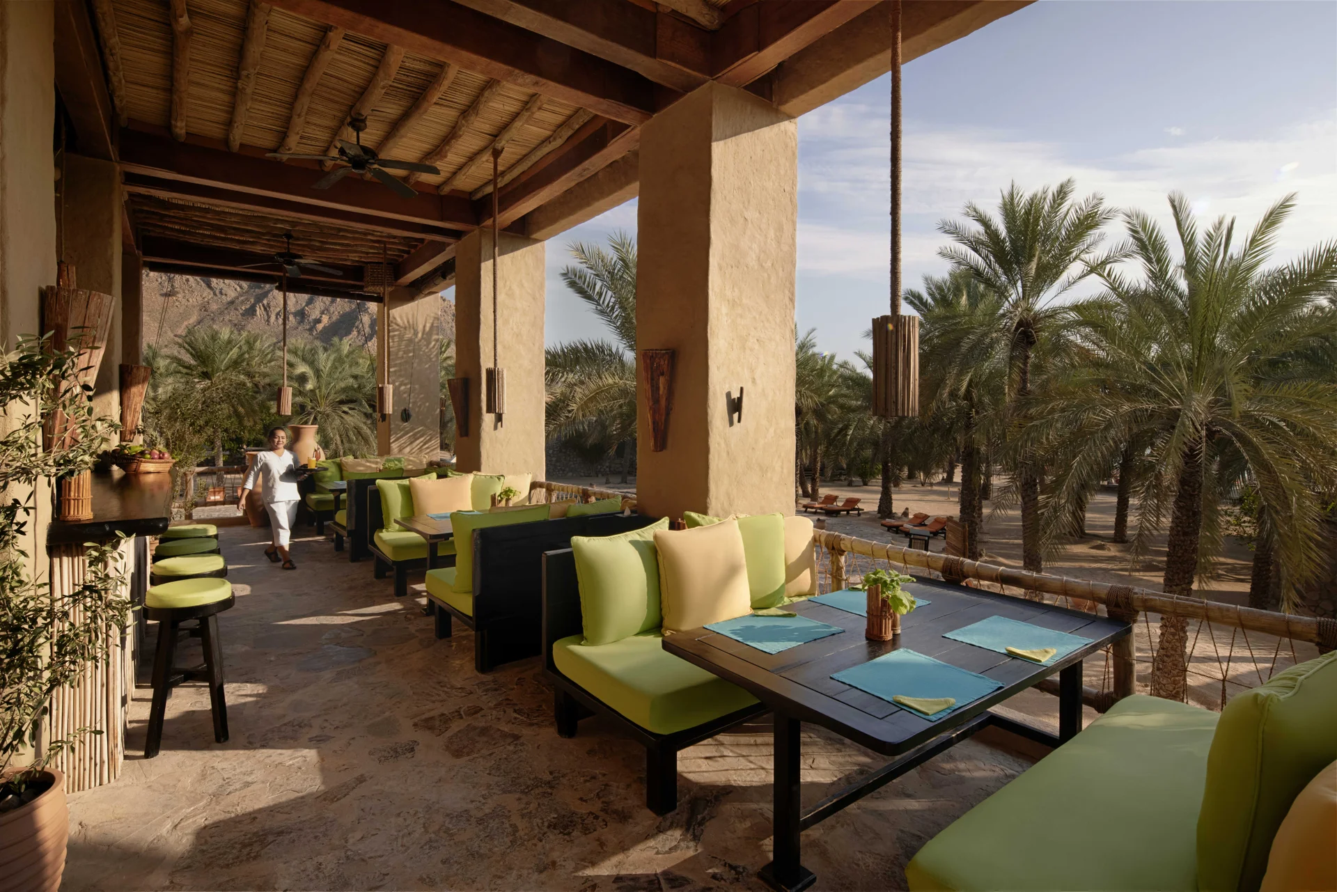 Zighy Bar's shaded terrace under a cane and timber roof, lime-green banquettes at dark tables laid with turquoise mats, looking out across a date palm grove to the mountains.