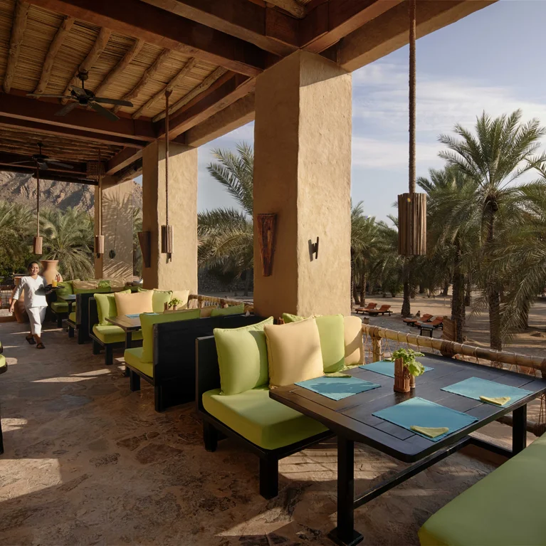 شرفة المطعم في الهواء الطلق مع مقاعد مبطنة بالوسائد الخضراء وطاولات خشبية بين أشجار النخيل، مع شخص يمشي في الخلفية.