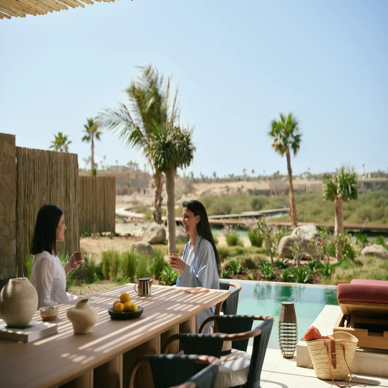 Guests enjoy refreshments at an outdoor table by a pool, surrounded by palm trees with desert hills in the distance.