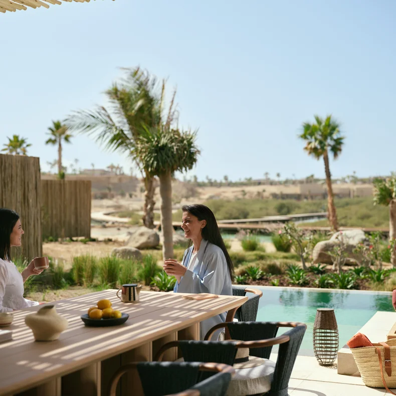 Guests enjoy refreshments at an outdoor table by a pool, surrounded by palm trees with desert hills in the distance.