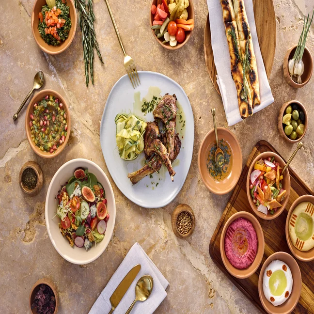 Mediterranean-inspired meal featuring grilled lamb chops, fresh salad, flatbread, assorted dips, pickled vegetables, olives and gold utensils on marble.