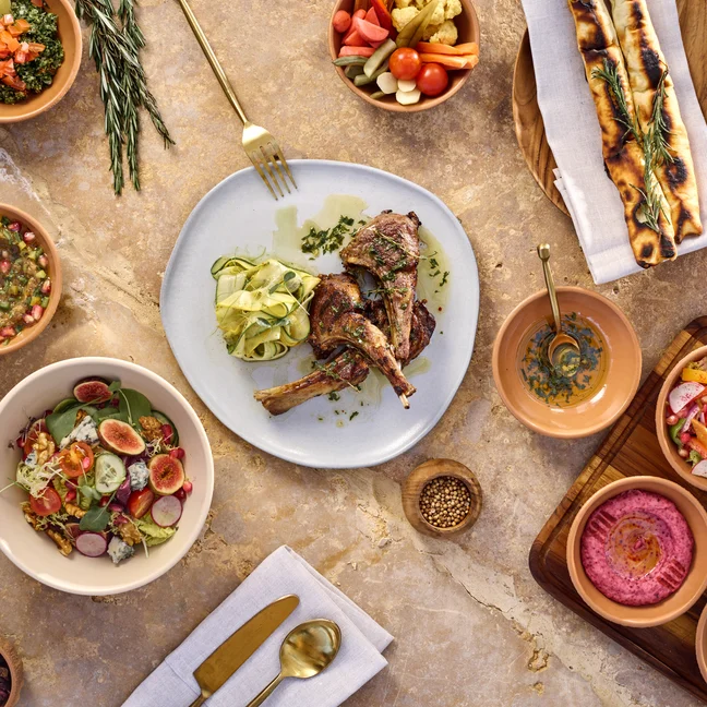 Mediterranean-inspired meal featuring grilled lamb chops, fresh salad, flatbread, assorted dips, pickled vegetables, olives and gold utensils on marble.