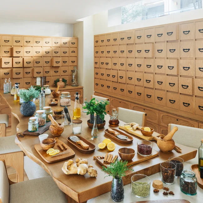Spacious room featuring wood cabinetry and a large table arranged with herbs, spices, glass bottles, and kitchen essentials for holistic wellness experiences.