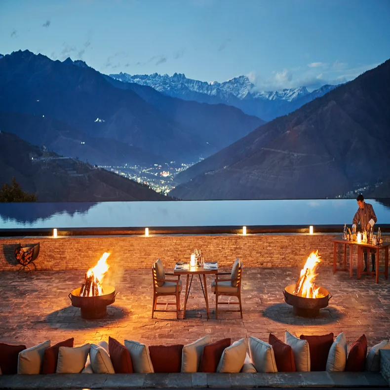 Dining terrace with fire features, arranged seating, and tableware faces mountains and valley at dusk; soft city lights dot the landscape in the distance.