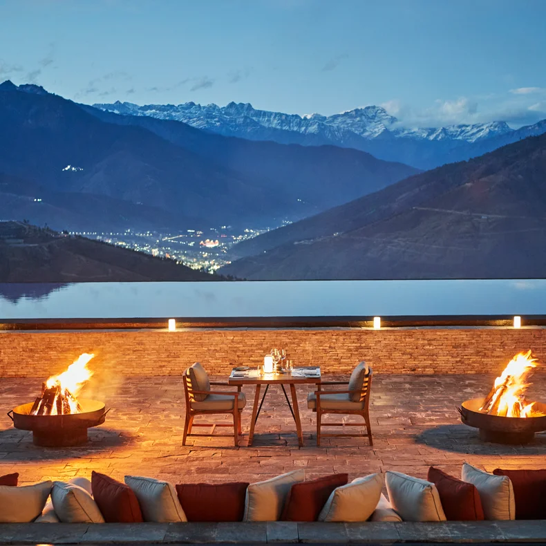Dining terrace with fire features, arranged seating, and tableware faces mountains and valley at dusk; soft city lights dot the landscape in the distance.