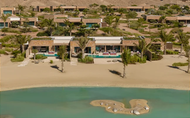 Villas with private pools sit along a sandy shore by a turquoise lagoon, set between desert landscapes and areas of abundant greenery.