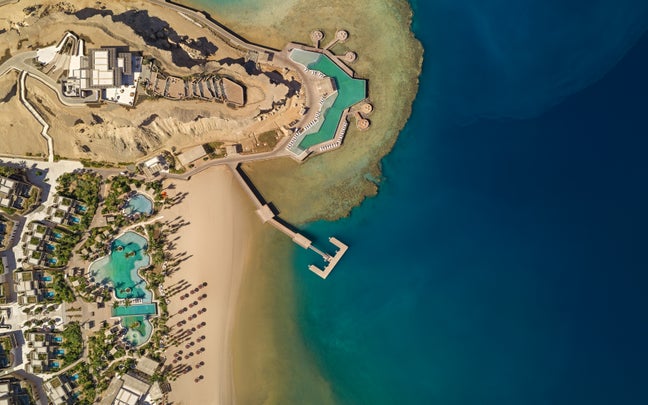 View of a beachfront resort featuring pools, sandy beach, a wooden pier, blue sea, and surrounding palm trees near modern low-rise buildings.