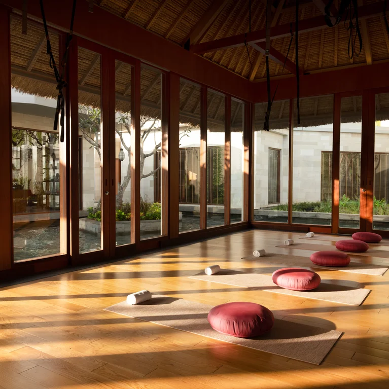 瑜伽室有大窗户、天然木地板、红色坐垫,垫子上铺有卷曲的毛巾。阳光洒满整个空间,营造出宁静的练习环境。.