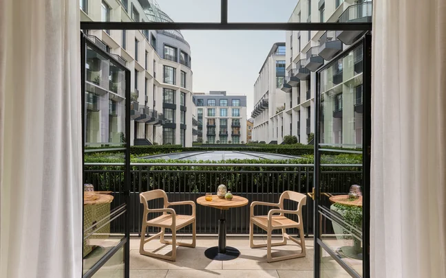 Two wooden chairs and a small round table on a balcony offer views of a green courtyard framed by contemporary buildings.