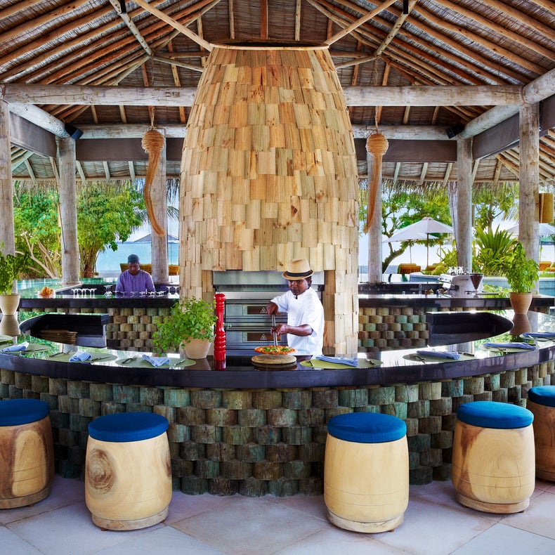 A thatched-roof bar with a curved counter and vibrant stools overlooks a pool, with chefs preparing dishes amid greenery.