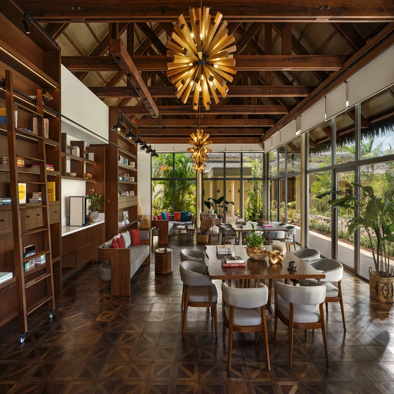 Open-plan room with exposed wood beams, tall windows for natural light, contemporary lighting, bookshelves, comfortable seating, dining area, and views of surrounding greenery.