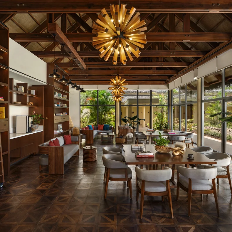 Open-plan room with exposed wood beams, tall windows for natural light, contemporary lighting, bookshelves, comfortable seating, dining area, and views of surrounding greenery.