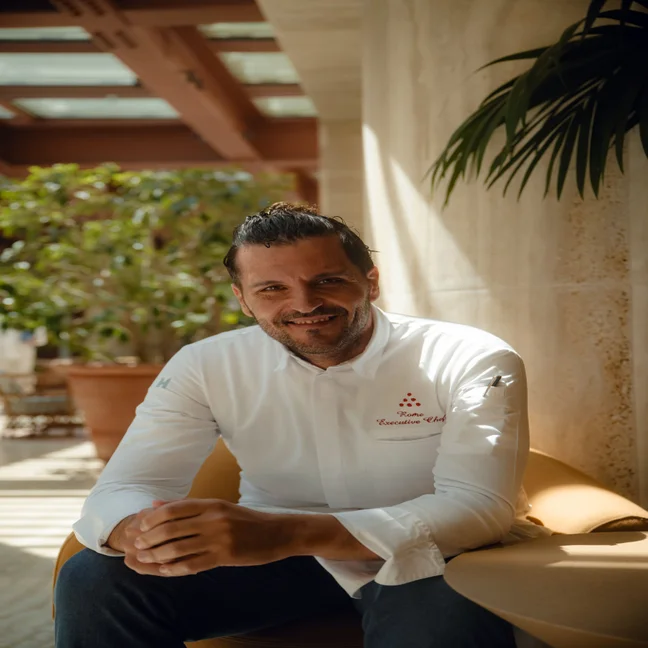 A chef in a white coat sits in a bright indoor setting with greenery, looking at the camera with a calm expression.