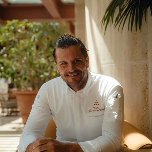 A chef in a white coat sits in a bright indoor setting with greenery, looking at the camera with a calm expression.