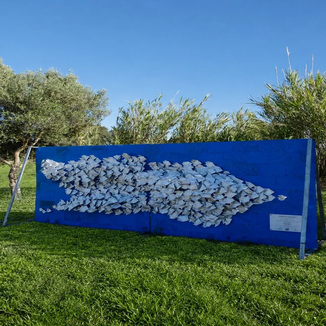 Blue outdoor art installation shaped like a fish with white folded elements, displayed on grass amidst trees and tall plants in a natural setting.