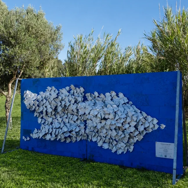 Blue outdoor art installation shaped like a fish with white folded elements, displayed on grass amidst trees and tall plants in a natural setting.