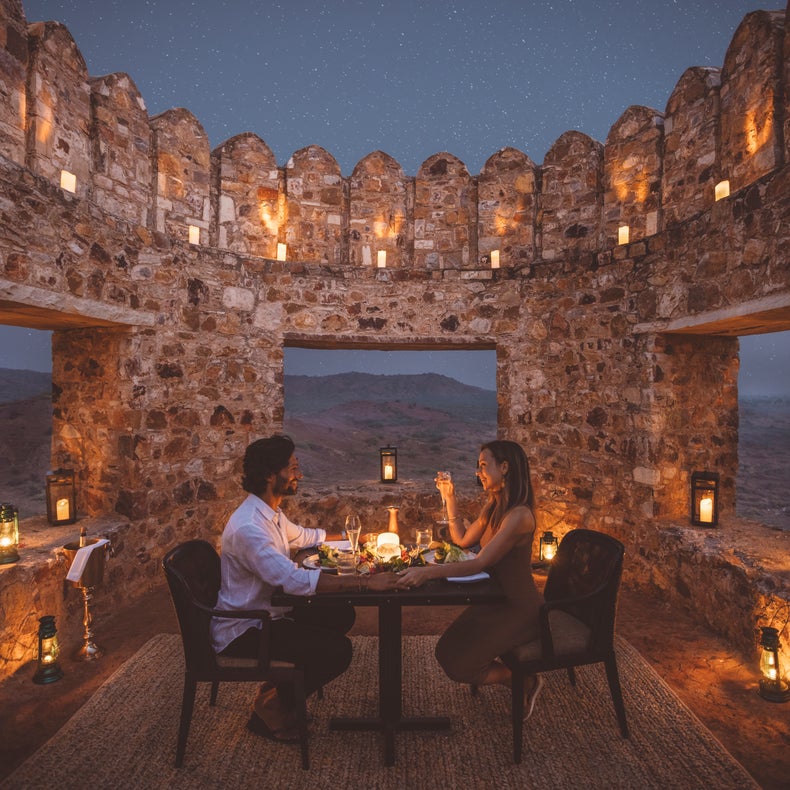 Los invitados disfrutan de una cena íntima a la luz de las velas en un entorno histórico de piedra con farolillos que iluminan las vistas de la montaña bajo un cielo despejado al atardecer.