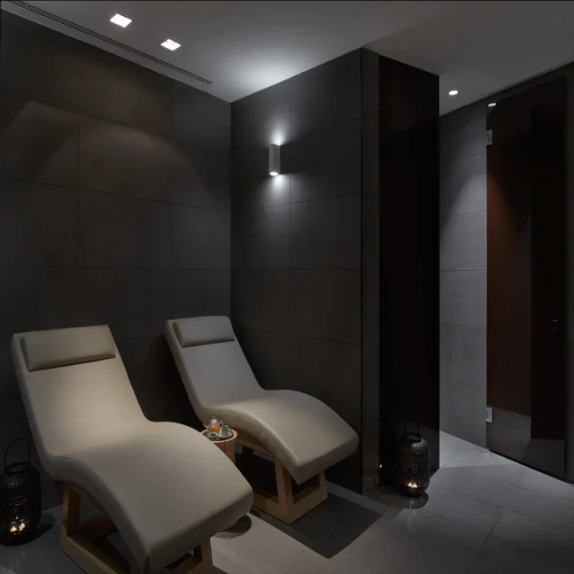 Beige lounge chairs in a softly lit spa space with dark tiled walls, a tray with tea cups set between them, and lanterns arranged on the floor for gentle ambiance.