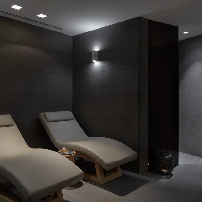 Beige lounge chairs in a softly lit spa space with dark tiled walls, a tray with tea cups set between them, and lanterns arranged on the floor for gentle ambiance.