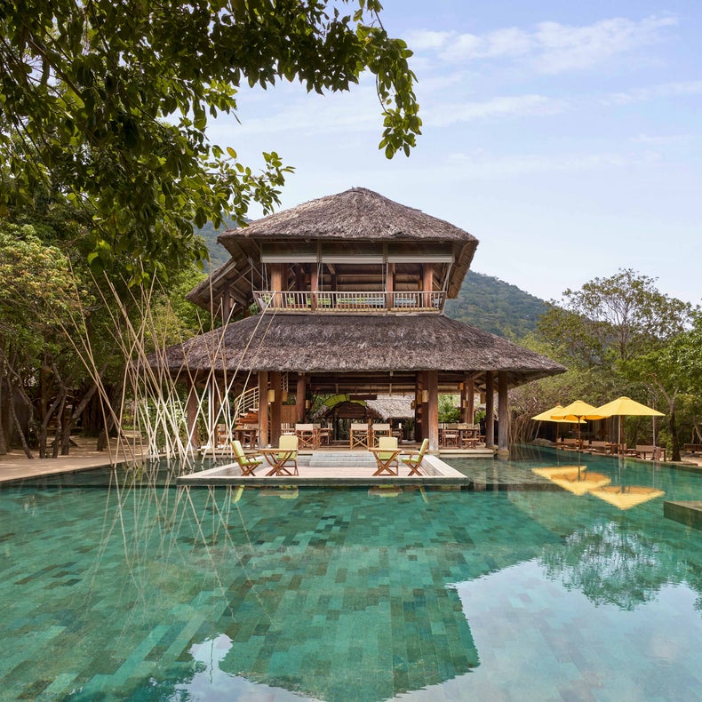 Un belvédère en bois au toit de chaume avec des parasols jaunes ombrage le ciel au bord de la piscine aux eaux cristallines, entourée d'une végétation luxuriante.