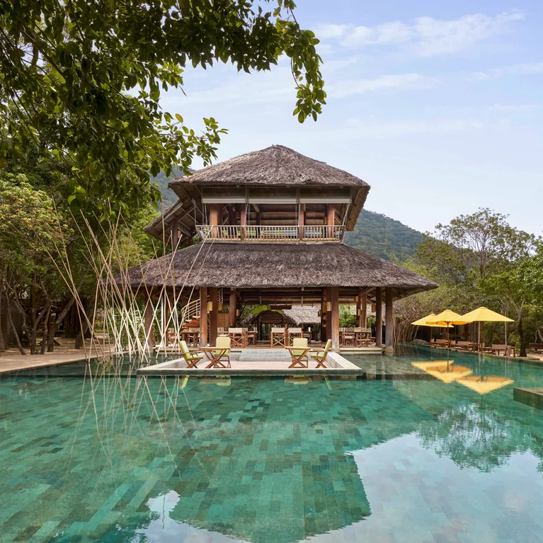 Bên cạnh bể bơi trong vắt là một ngôi nhà gỗ mái tranh, những chiếc ô che nắng màu vàng che phủ bầu trời, xung quanh là những hàng cây xanh mát.