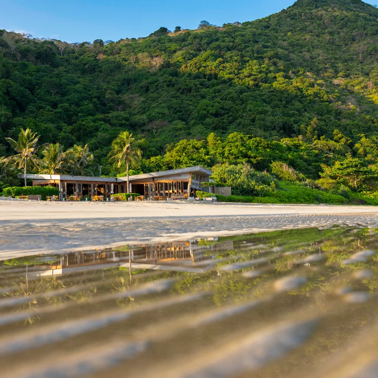 A thoughtfully designed beachside retreat set beside green hills, with palm trees and clear shallow water gently reflecting the landscape on a sandy shore.