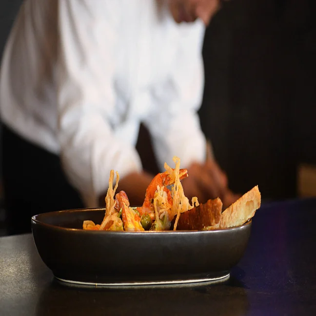 A plated dish with shrimp, crisp toppings, and bread sits in focus on a dark plate, as a chef works in the softly blurred background.