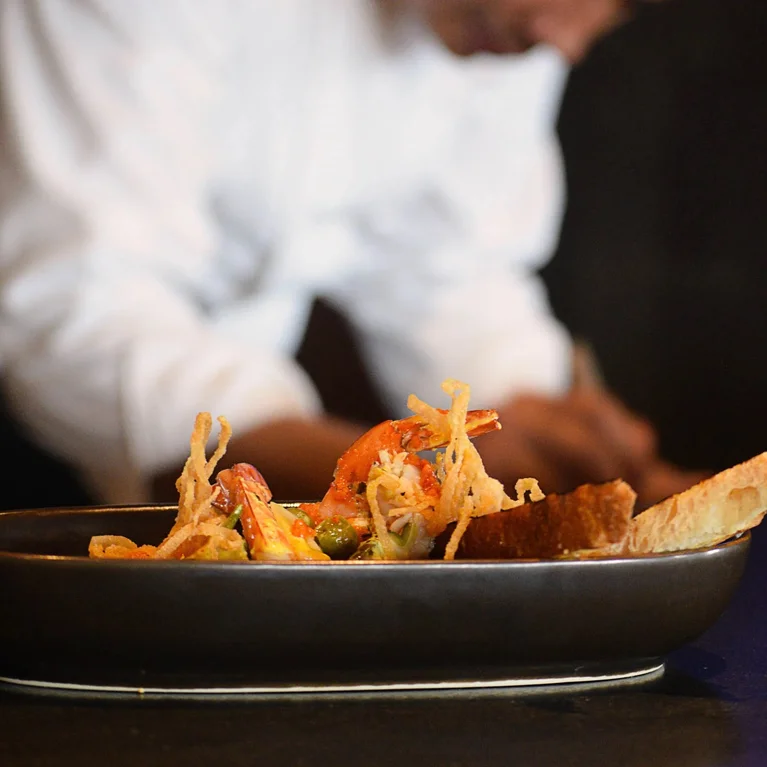 طبق مطهو مع الروبيان والطبقة المقرمشة والخبز في طبق داكن، بينما يعمل الطاهي في الخلفية الضبابية الهادئة.