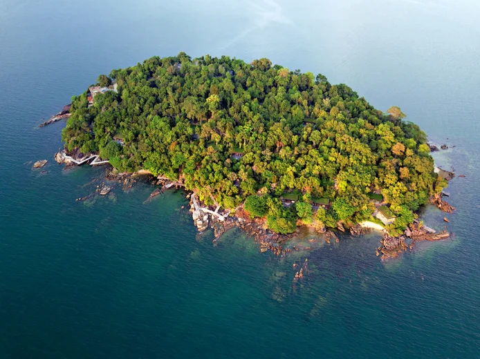 An island with vibrant greenery and dense trees, surrounded by tranquil blue ocean and natural rocky shoreline, seen from an overhead perspective.